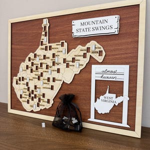 May include: A rectangular wooden wall plaque featuring a light-colored laser-cut map of West Virginia with numerous small rectangular slots. The text reads MOUNTAIN STATE SWINGS and almost heaven WEST VIRGINIA. A black mesh drawstring bag holds small tokens.