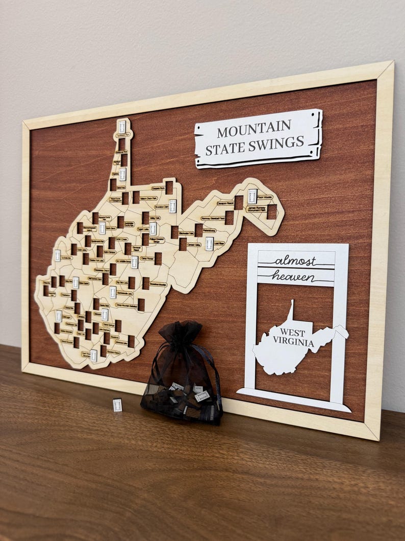 May include: A rectangular wooden wall plaque featuring a light-colored laser-cut map of West Virginia with numerous small rectangular slots. The text reads MOUNTAIN STATE SWINGS and almost heaven WEST VIRGINIA. A black mesh drawstring bag holds small tokens.