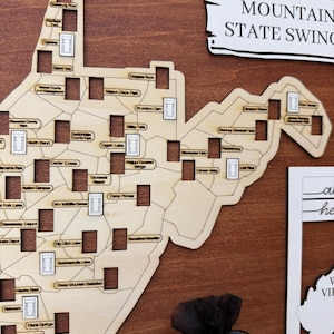 May include: A wooden map of West Virginia featuring laser-cut outlines and rectangular slots for tokens. The map includes text labels for various locations such as Tygart Lake and Canaan Valley, with a sign reading MOUNTAIN STATE SWING.