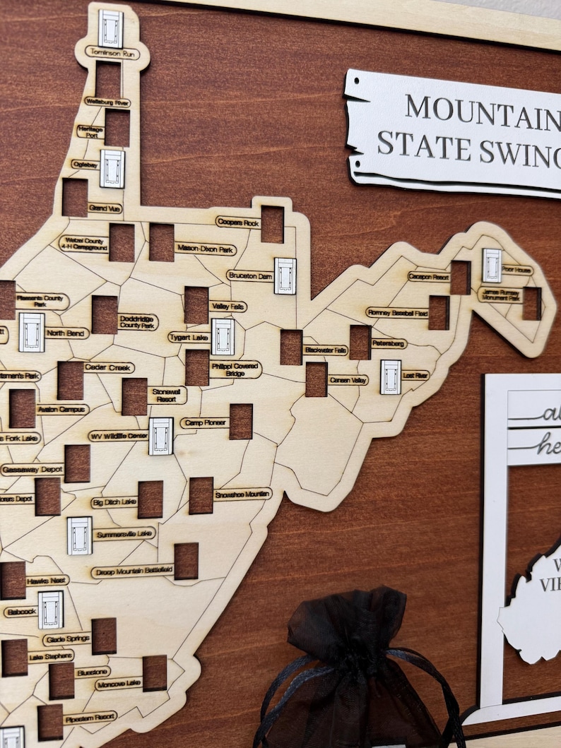 May include: A wooden map of West Virginia featuring laser-cut outlines and rectangular slots for tokens. The map includes text labels for various locations such as Tygart Lake and Canaan Valley, with a sign reading MOUNTAIN STATE SWING.