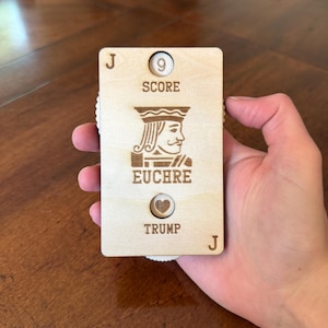 May include: A wooden Euchre scorekeeper in the shape of a playing card, held in a hand. The card features the Jack of Hearts, the word "SCORE" with the number 9, and the words "EUCHRE" and "TRUMP". The scorekeeper has a rotating dial to track the score.