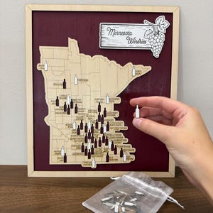 May include: A wooden map of Minnesota featuring labeled wineries, mounted on a dark red background within a light wood frame. A sign at the top reads Minnesota Wineries with a grape cluster illustration. Small wooden wine bottle markers are included.