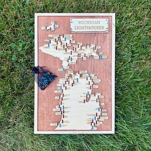 May include: Wooden map of Michigan with lighthouse markers. The map is framed in light wood and features the text "MICHIGAN LIGHTHOUSES" at the top. Small wooden lighthouse markers are placed throughout the map, with a small bag of blue and black markers in the corner.