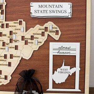 May include: A wooden wall map of West Virginia featuring laser-cut details, a sign reading MOUNTAIN STATE SWINGS, and a white frame with the text almost heaven WEST VIRGINIA. A black organza drawstring bag contains small wooden tokens.
