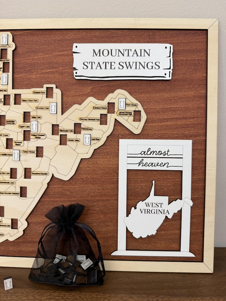 May include: A wooden wall map of West Virginia featuring laser-cut details, a sign reading MOUNTAIN STATE SWINGS, and a white frame with the text almost heaven WEST VIRGINIA. A black organza drawstring bag contains small wooden tokens.