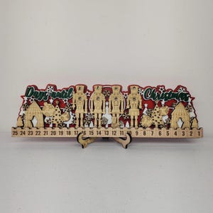 May include: Wooden Christmas advent calendar with a red and green design. The calendar features laser-cut nutcrackers, houses, and Christmas trees. The words "Days until Christmas" are displayed across the top, with numbers 1-25 below.