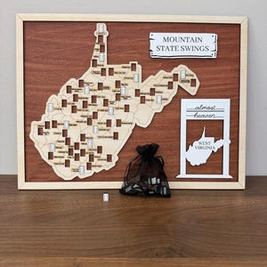 May include: A wooden wall map of West Virginia featuring cutouts for swing locations, a sign reading MOUNTAIN STATE SWINGS, and a white frame with the text almost heaven WEST VIRGINIA. A black organza bag contains small wooden swing tokens.