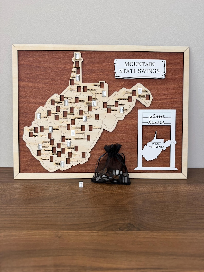 May include: A wooden wall map of West Virginia featuring cutouts for swing locations, a sign reading MOUNTAIN STATE SWINGS, and a white frame with the text almost heaven WEST VIRGINIA. A black organza bag contains small wooden swing tokens.