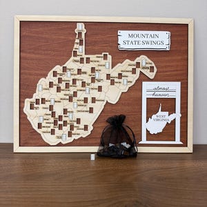 May include: A wooden map of West Virginia featuring cutouts for swing locations, framed in light wood. It includes a sign reading MOUNTAIN STATE SWINGS, a white sign with the text almost heaven WEST VIRGINIA, and a black mesh bag containing small tokens.
