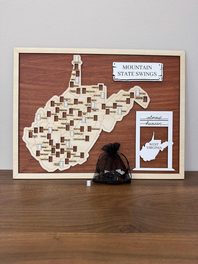 May include: A wooden map of West Virginia featuring cutouts for swing locations, framed in light wood. It includes a sign reading MOUNTAIN STATE SWINGS, a white sign with the text almost heaven WEST VIRGINIA, and a black mesh bag containing small tokens.