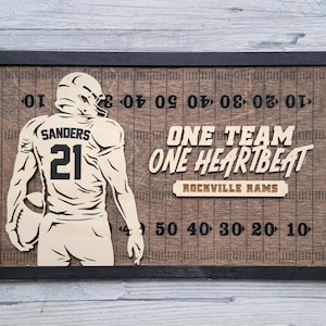 May include: A wooden sign with a black frame depicting a football player in a jersey with the number 21 and the name "Sanders" on it. The sign has a football field background with yard lines and the text "One Team One Heartbeat Rockville Rams".