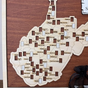 May include: A wooden wall map of West Virginia featuring a light wood cutout of the state shape mounted on a dark wood background. The map includes numerous rectangular cutouts labeled with various state locations and a black mesh drawstring bag.