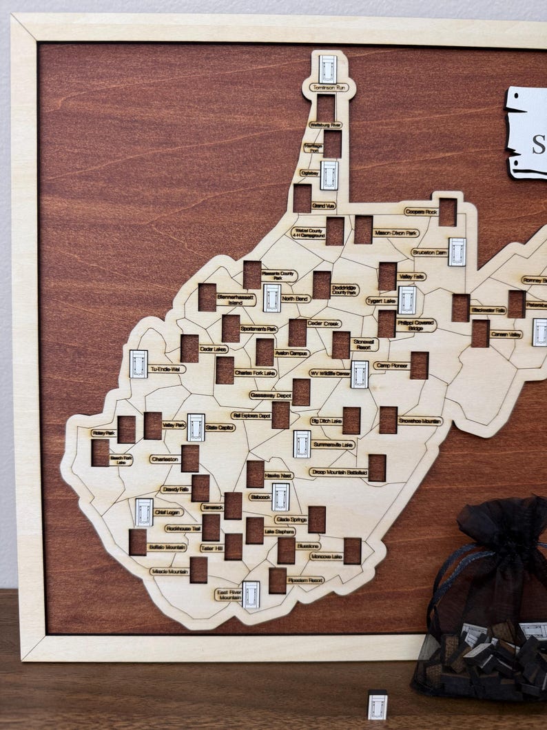 May include: A wooden wall map of West Virginia featuring a light wood cutout of the state shape mounted on a dark wood background. The map includes numerous rectangular cutouts labeled with various state locations and a black mesh drawstring bag.