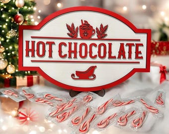 May include: A red and white wooden sign that reads "HOT CHOCOLATE" with a cup of hot chocolate and a sleigh design. Candy canes are scattered in front of the sign, with a Christmas tree in the background. The sign is a festive decoration.