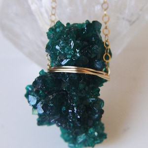 May include: A dark green crystal pendant necklace with a gold chain. The crystal is a cluster of small, sparkling formations. The pendant is held in place by a gold wire. The necklace is displayed on a white surface.