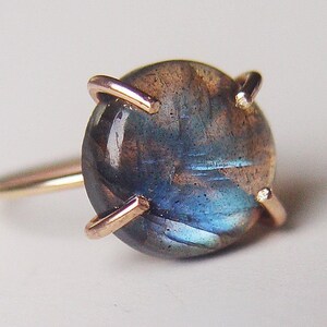 Close-up of a rose gold ring featuring a cabochon-cut labradorite gemstone. The dark blue-green stone displays iridescent flashes.  The setting is a simple, four-prong design with delicate rose gold wire. The ring showcases a minimalist aesthetic, highlighting the gemstone's natural beauty.