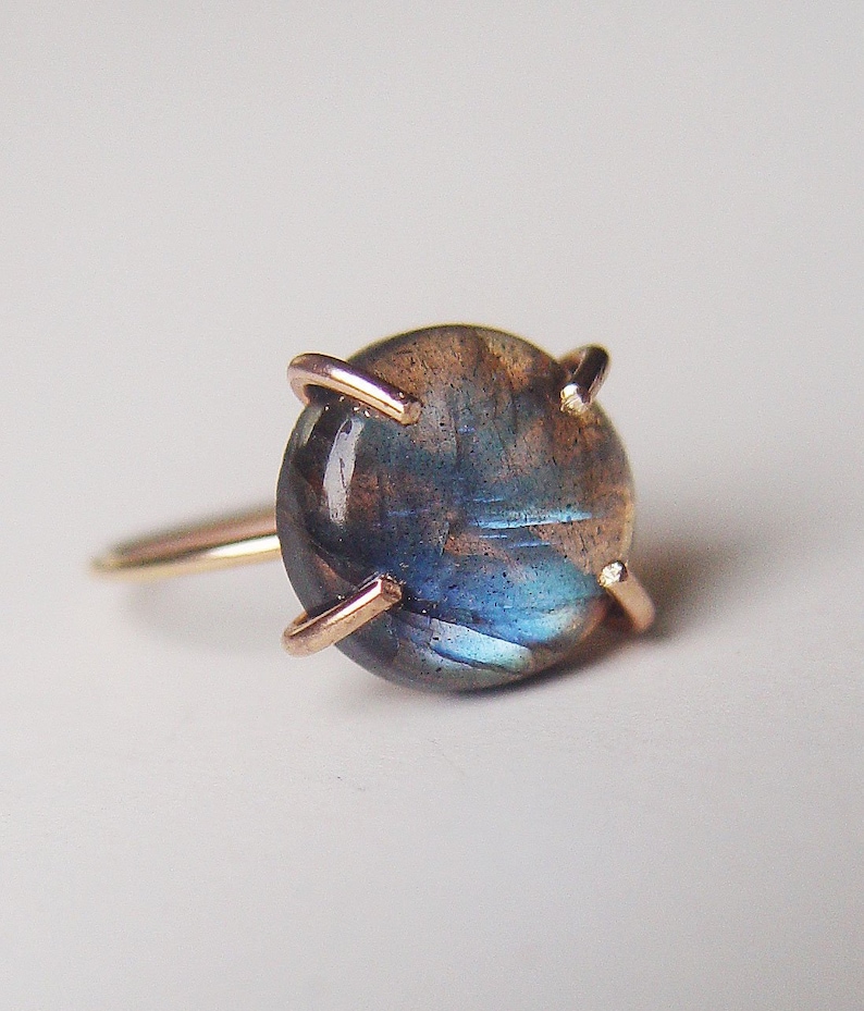 Close-up of a rose gold ring featuring a cabochon-cut labradorite gemstone. The dark blue-green stone displays iridescent flashes.  The setting is a simple, four-prong design with delicate rose gold wire. The ring showcases a minimalist aesthetic, highlighting the gemstone's natural beauty.