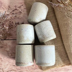 Peut inclure: Cinq tasses en céramique tachetée de différentes nuances de blanc et de beige sont disposées sur une surface en bois avec une toile de jute. Les tasses ont une forme cylindrique avec une base arrondie, parfaites pour les boissons.