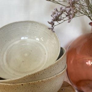 Peut inclure: Trois bols en céramique blanche avec une texture mouchetée. Les bols sont empilés les uns sur les autres. Le bol du haut est lisse et les deux bols du bas ont une texture mouchetée.