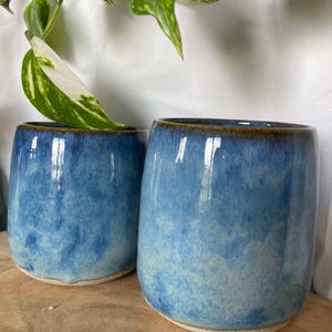 Peut inclure: Deux pots en céramique bleus avec un glaçage blanc et un bord brun. Les pots sont posés sur une surface en bois.
