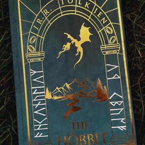 May include: A teal hardcover book titled "The Hobbit" by J.R.R. Tolkien. The cover features gold foil accents, including a dragon, mountains, and stylized text. The book is set against a backdrop of green grass.