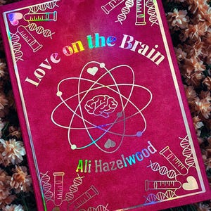 Puede incluir: Un libro de terciopelo rojo con una cubierta holográfica que presenta el título "Love on the Brain" y el nombre del autor "Ali Hazelwood". La cubierta también incluye un gráfico de un átomo con un cerebro en su interior y hebras de ADN.