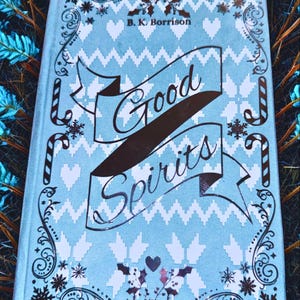 May include: A light blue book cover with the title "Good Spirits" in a banner. The cover has a repeating pattern of white hearts and zigzags, with decorative snowflakes and candy canes. The author's name, B. K. Borrison, is at the top.