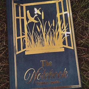 May include: A hardcover book titled "The Notebook" by Nicholas Sparks. The cover is navy blue with gold foil accents, featuring a window design with flying ducks and cattails. The book is resting on a bed of green grass.
