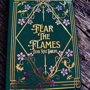 May include: A hardcover book with a dark green cover featuring gold and pink floral and bat designs. The title "Fear the Flames" is in gold lettering, with the author's name below. The book has gold border accents.