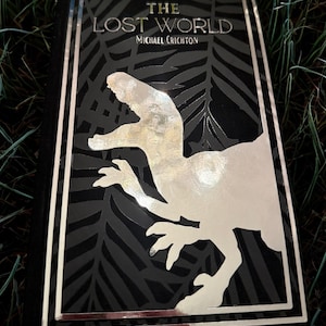 May include: Hardcover book titled "The Lost World" by Michael Crichton. The cover is black with a silver dinosaur illustration and text. The book has a silver border and is set against a background of green grass.