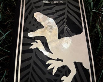 May include: Hardcover book titled "The Lost World" by Michael Crichton. The cover is black with a silver dinosaur illustration and text. The book has a silver border and is set against a background of green grass.