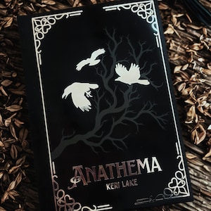 Könnte beinhalten: Ein schwarzes gebundenes Buch mit dem Titel Anathema von Keri Lake. Der Einband zeigt einen metallisch goldenen Rand mit Eckverzierungen, die Silhouette eines kahlen Baumes und drei weiße Vögel im Flug. Titel und Autor sind in Gold gedruckt.