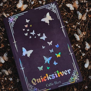 Puede incluir: Un libro de tapa dura de color morado oscuro con un diseño de mariposas en lámina plateada y el título "Quicksilver" de Callie Hart.