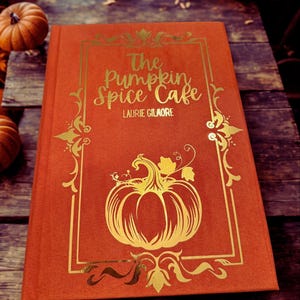 Op de afbeelding: Een oranje boek met de titel "The Pumpkin Spice Cafe" met gouden letters en een pompoenillustratie. Het boek ligt op een houten oppervlak, met pompoenen op de achtergrond, wat een herfstthema oproept.