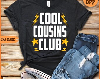May include: A black t-shirt with the text "Cool Cousins Club" in white letters with yellow lightning bolts and stars. The shirt is laid out on a wooden surface with a pair of blue jeans and white sneakers.