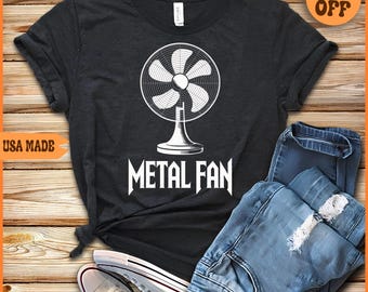 Può includere: T-shirt grigio scuro con grafica bianca di un ventilatore e la scritta "METAL FAN". La maglietta è abbinata a jeans blu e scarpe da ginnastica bianche. Un adesivo indica "20% OFF".
