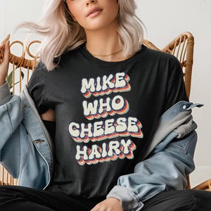 May include: A black t-shirt with a retro-style graphic that says "Mike Who Cheese Hairy".