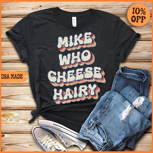 May include: A dark gray t-shirt with a retro-style graphic that says "Mike Who Cheese Hairy". The shirt is laid out on a wooden surface with a pair of white sneakers and blue jeans.