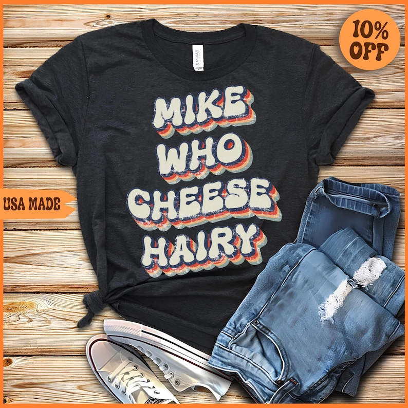 May include: A dark gray t-shirt with a retro-style graphic that says "Mike Who Cheese Hairy". The shirt is laid out on a wooden surface with a pair of white sneakers and blue jeans.