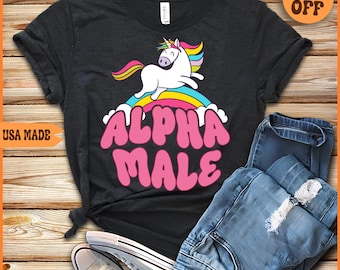 May include: Black t-shirt featuring a pink and white unicorn graphic and the text "Alpha Male" in pink letters. The unicorn is standing on a rainbow.