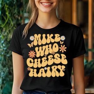 May include: Black t-shirt with a retro-style graphic that says "Mike Who Cheese Hairy" in yellow letters with flowers and butterflies.