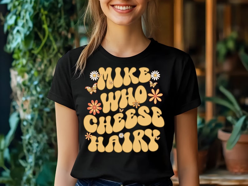 May include: Black t-shirt with a retro-style graphic that says "Mike Who Cheese Hairy" in yellow letters with flowers and butterflies.