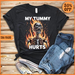May include: Dark gray t-shirt with the text "MY TUMMY HURTS" above a skeleton graphic engulfed in flames. The shirt is paired with blue jeans and white sneakers. A "USA MADE" label is visible.