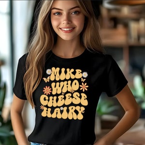 May include: Black t-shirt featuring a retro-style graphic that reads "Mike Who Cheese Hairy" in yellow lettering with orange and white flowers and butterflies.
