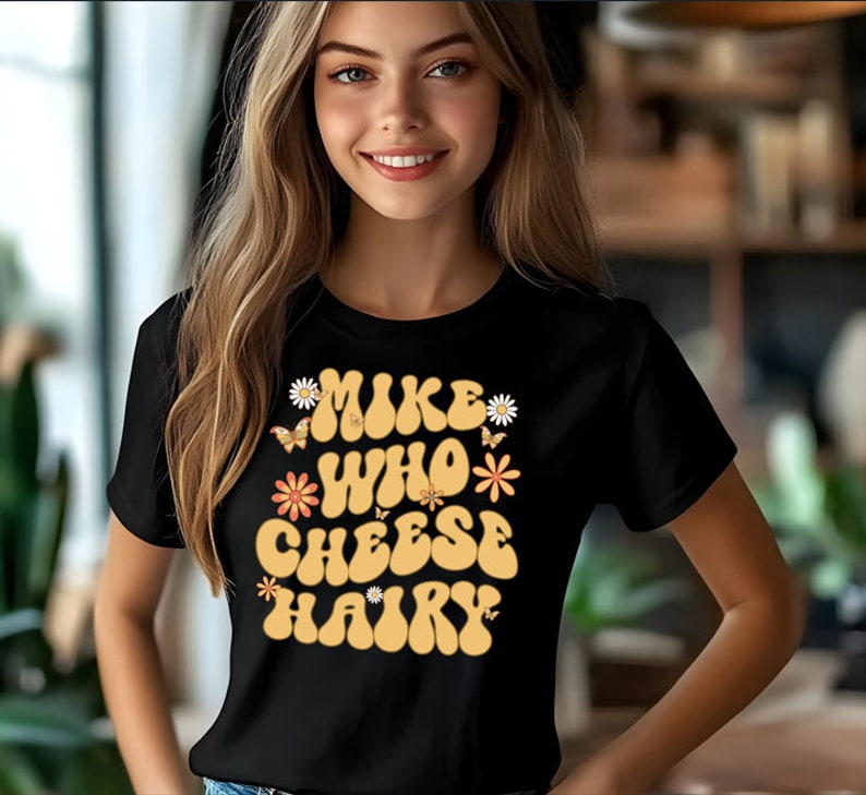 May include: Black t-shirt featuring a retro-style graphic that reads "Mike Who Cheese Hairy" in yellow lettering with orange and white flowers and butterflies.
