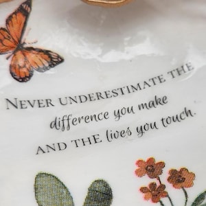 May include: A white decorative oyster shell dish with a gold-painted rim. The interior features an orange butterfly, small orange flowers, green foliage, and the black text: NEVER UNDERESTIMATE THE difference you make AND THE lives you touch.