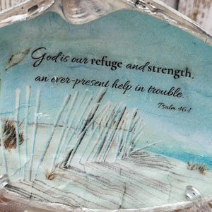 May include: A decorative seashell with a painted beach scene and the text "God is our refuge and strength, an ever-present help in trouble. Psalm 46:1." The shell has a silver rim and is displayed on a clear stand, surrounded by other shells.