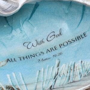 May include: A decorative seashell with a silver-colored rim. The interior features a blue and white beach scene with the text "With God, All Things Are Possible, Matthew 19:26."