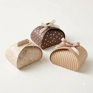 May include: Three curved gift boxes with ribbon bows. One is dark brown with gold polka dots and a cream ribbon. One is light beige with a white floral pattern. One is tan with white vertical stripes and a light brown satin ribbon bow on top.