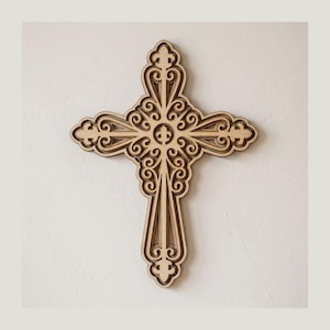 May include: A light-colored wooden wall cross featuring intricate laser-cut scrollwork and filigree patterns. The design includes a central fleur-de-lis motif and symmetrical, layered cutouts that create a detailed, openwork appearance.